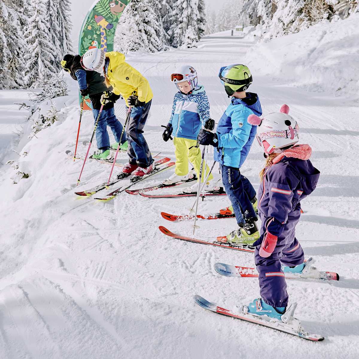 Kindergruppe auf Ski am Pistenrand, vorne ein Mädchen mit Hasenohren-Helm