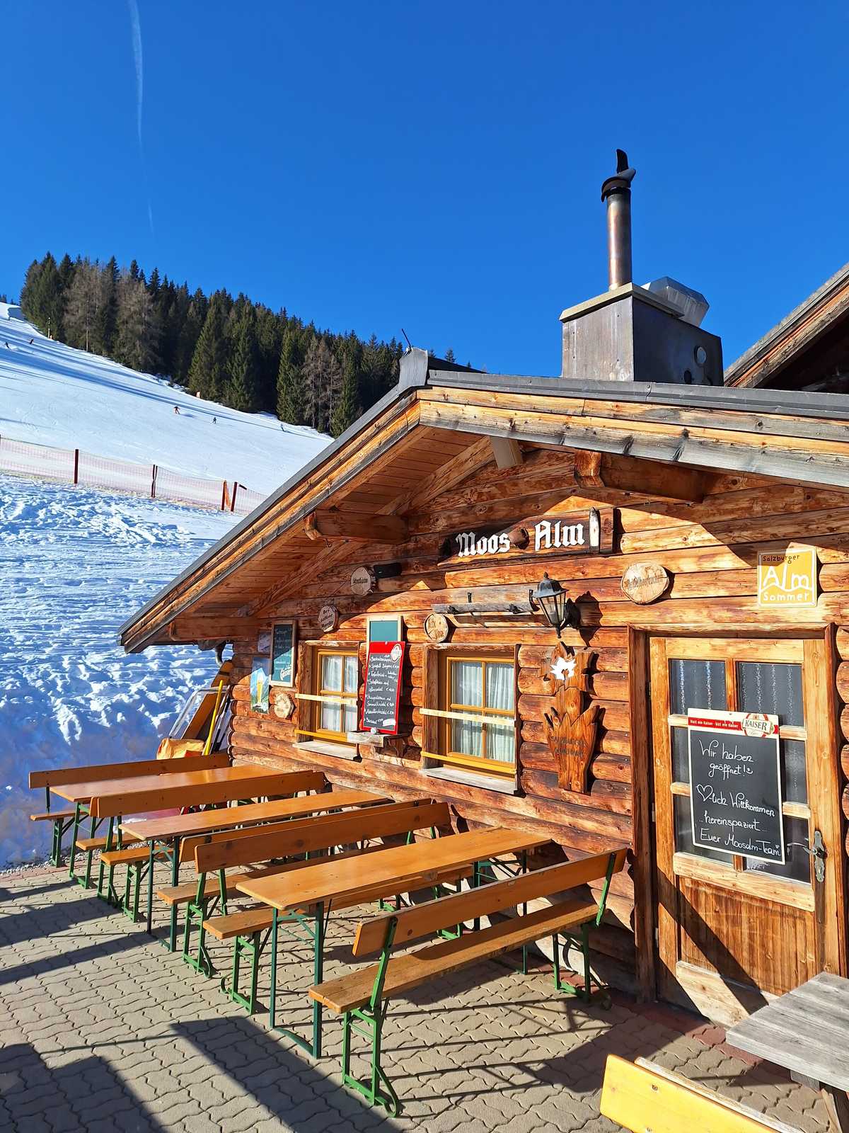 Die Moosalm mit Terrasse in der Wintersonne, links die Piste