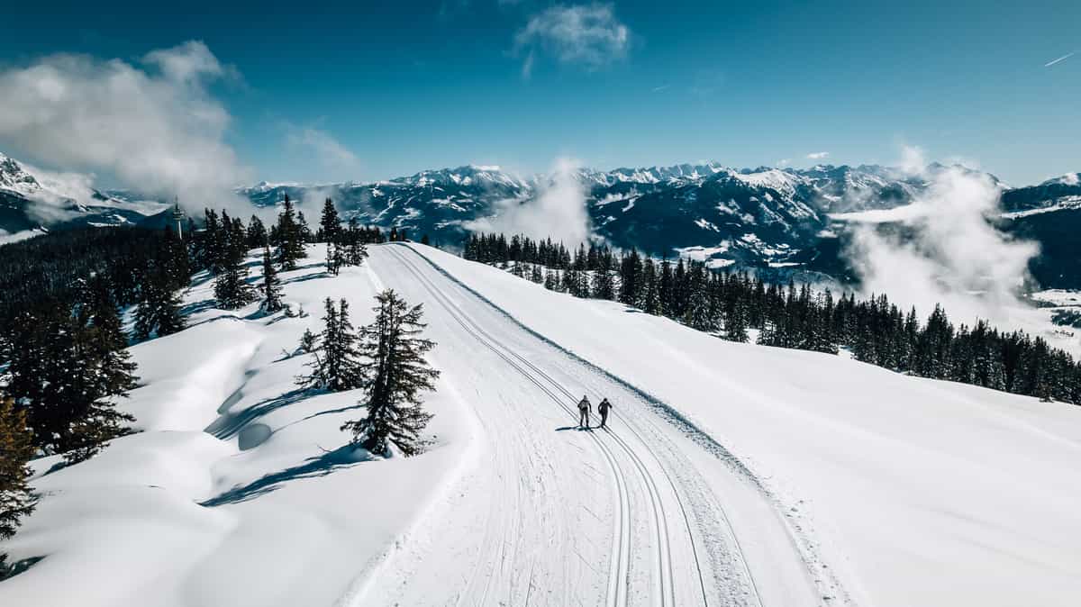 Luftbild: Loipe auf Bergrücken mit zwei Langläufern, Bergpanorama und Wolken