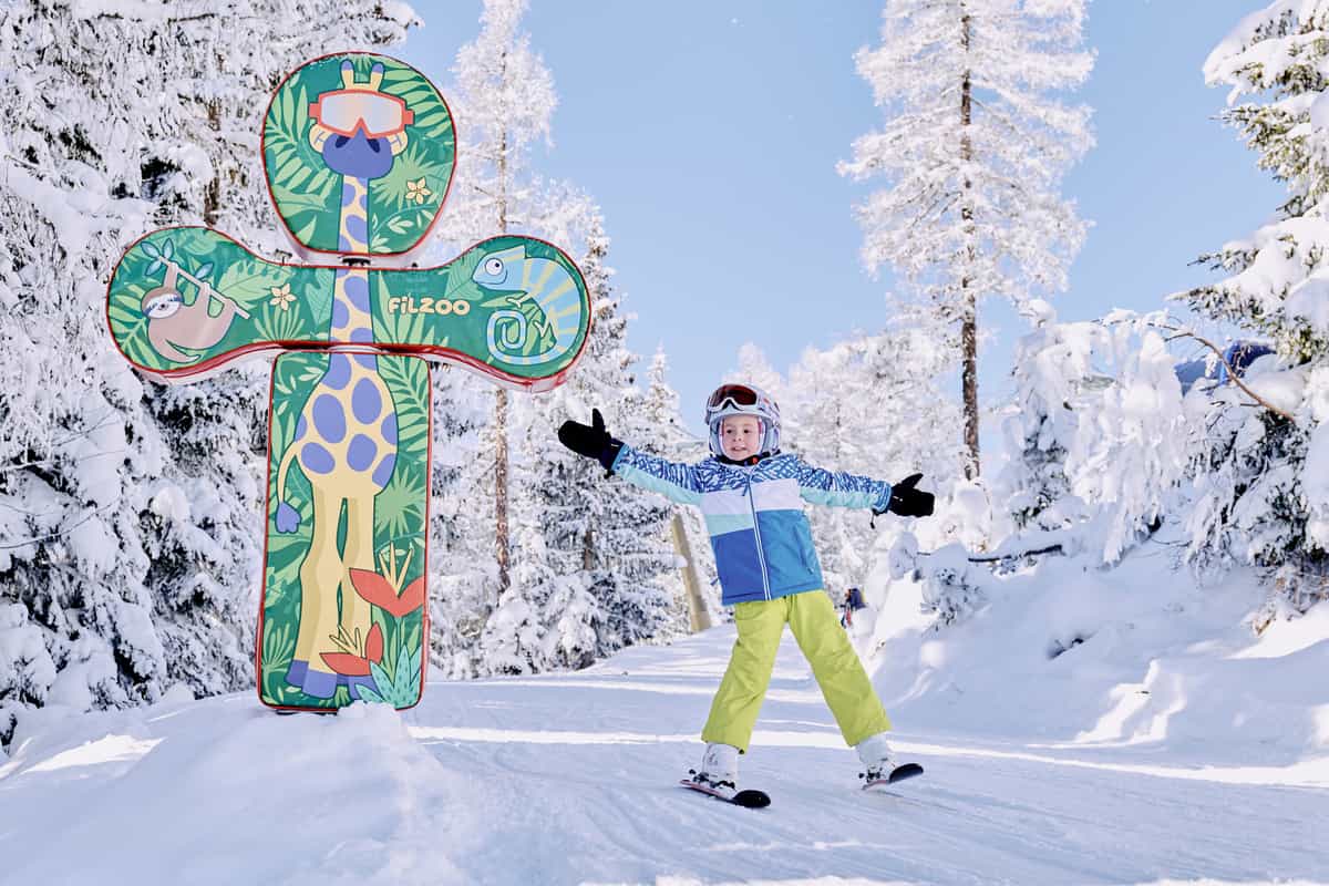 Kind auf Ski breitet vor der FiLZOO-Tafel die Arme aus, verschneiter Wald