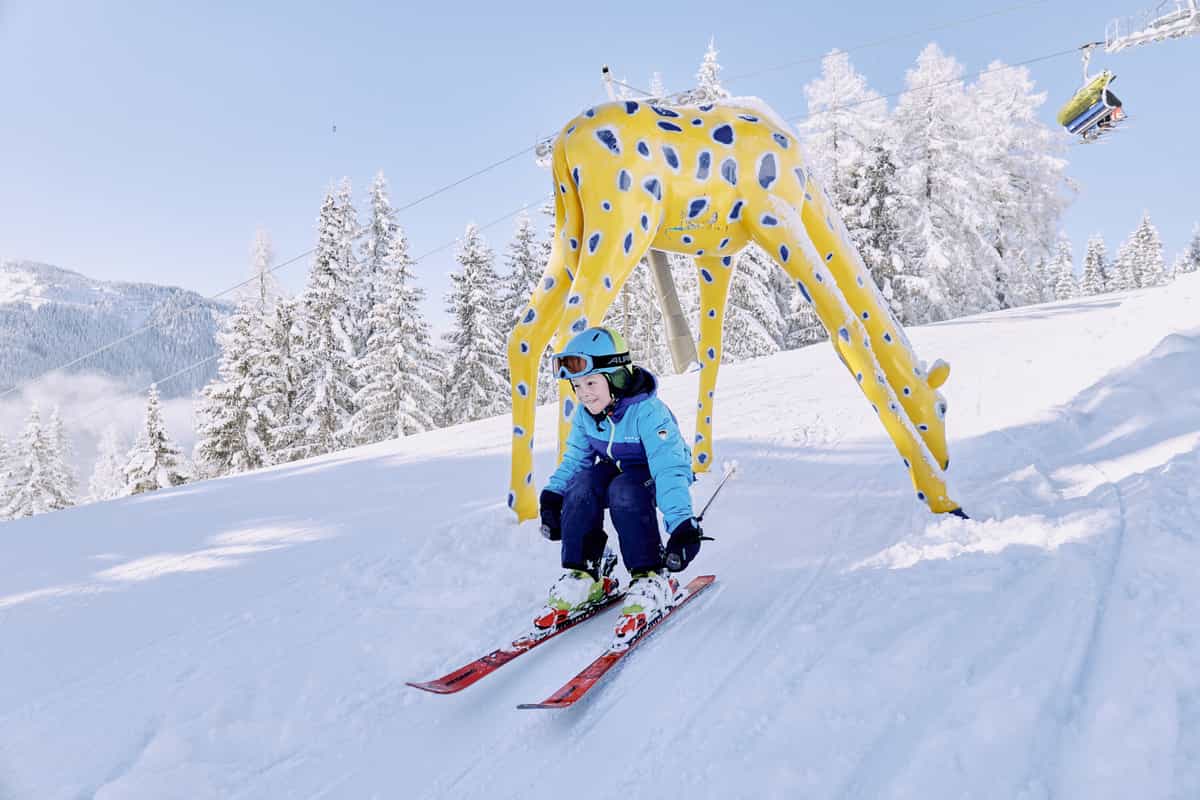 Kind auf Ski fährt unter dem Bogen der gelben Giraffe durch, Sessellift im Hintergrund