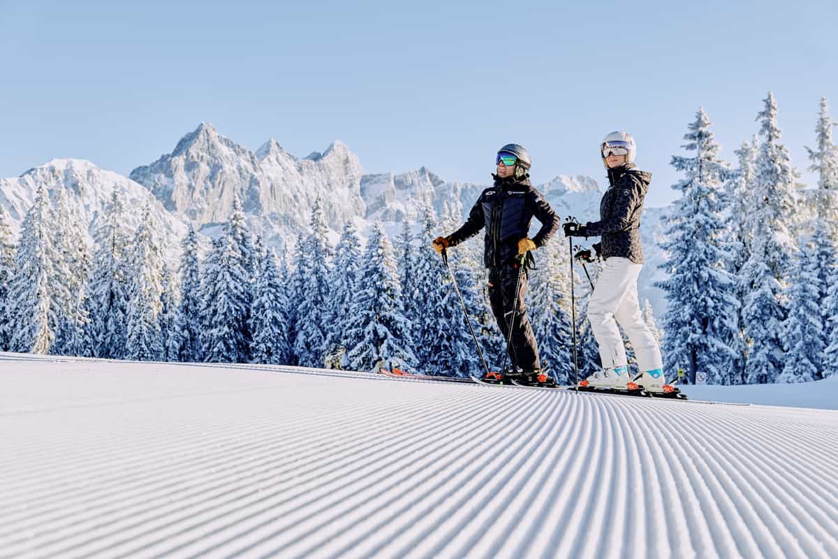 Paar klein auf weiter Cord-Piste, Felswand und Winterwald, viel Vordergrund