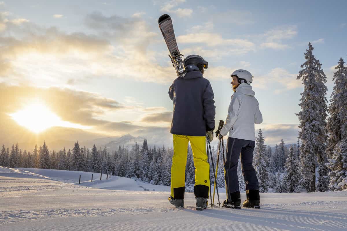 Skipaar mit geschulterten Skiern blickt in die tiefstehende Sonne über Winterwald