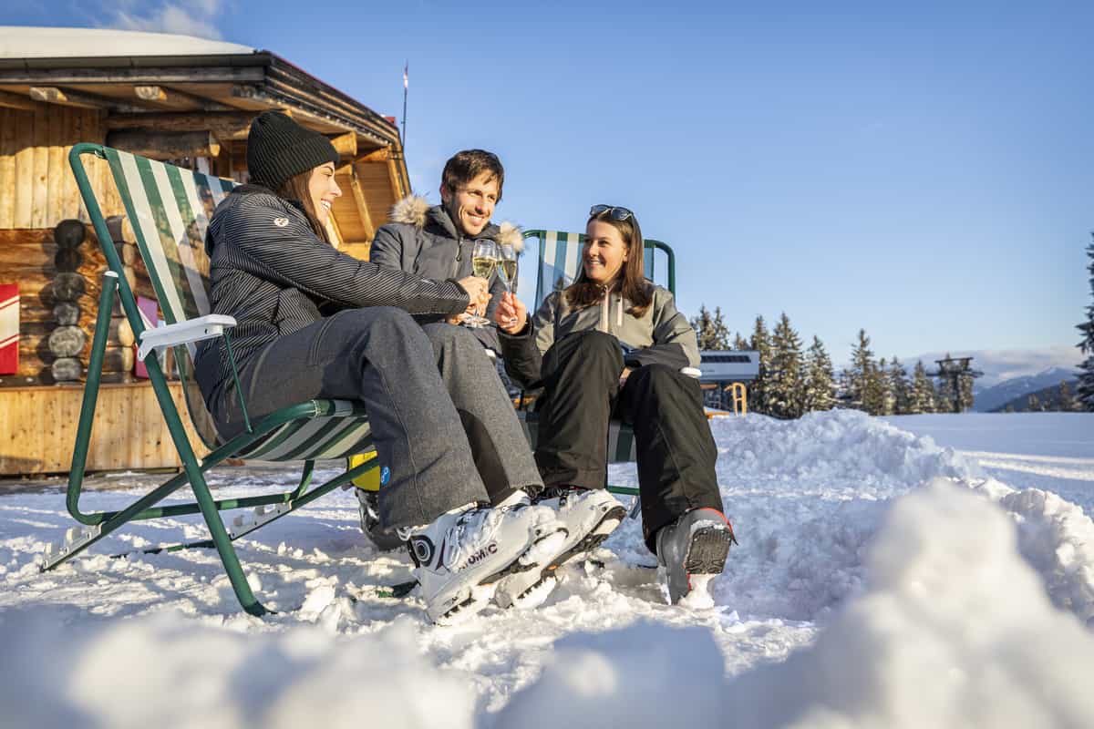 Drei Freunde im Liegestuhl vor der Holzhütte im Schnee, Sektgläser, blauer Himmel