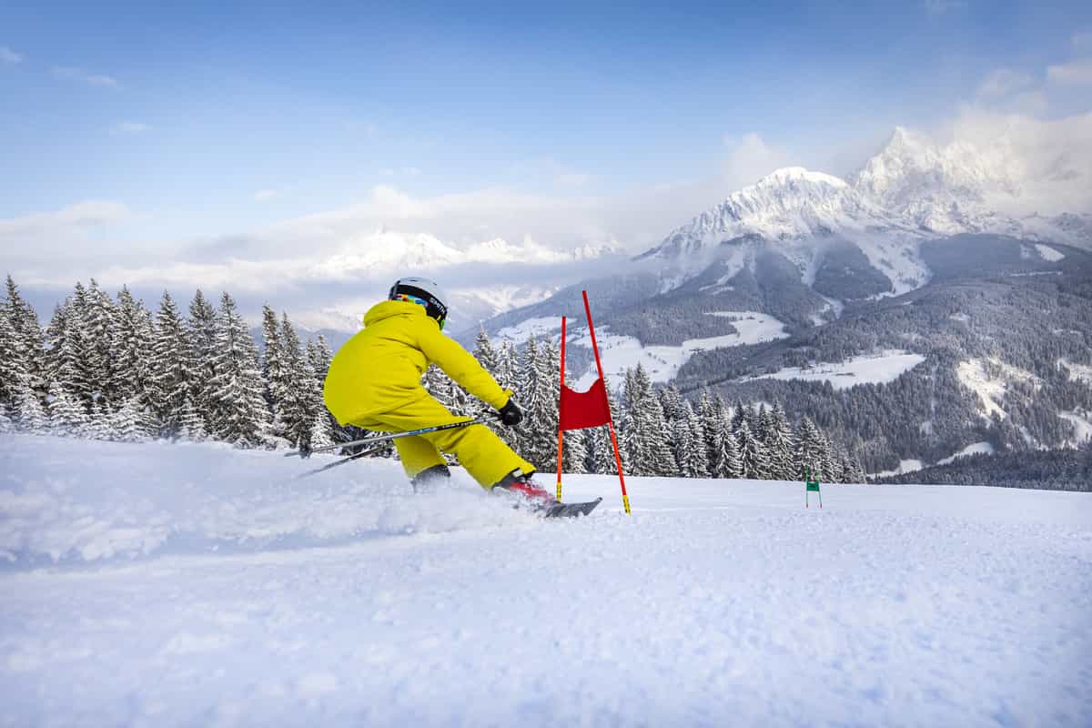 Kind in Gelb fährt am Tor vor Bergpanorama, Schnee spritzt